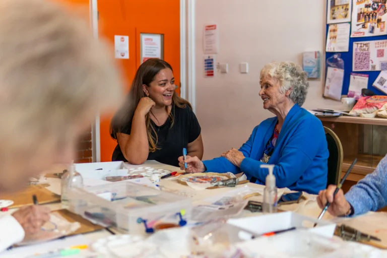An older woman smiling with another participant at a table covered in art materials