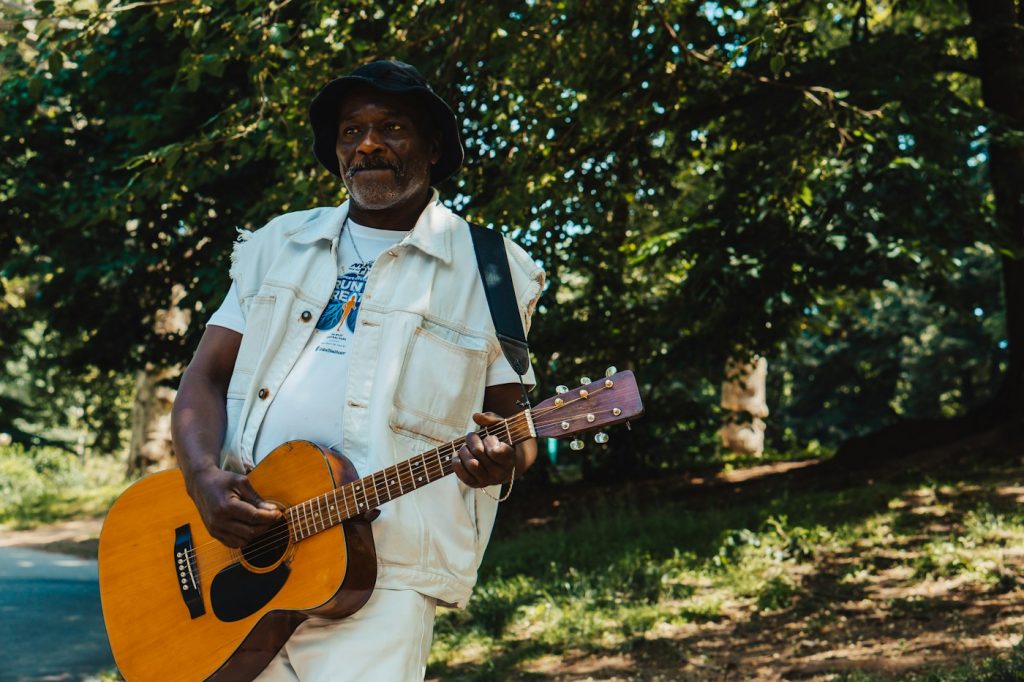 A musician holding an acoustic guitar beneath green trees in New York