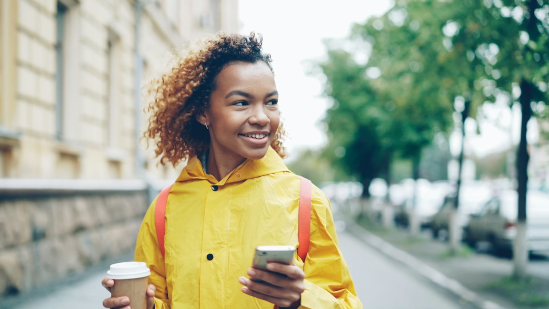 A smiling woman using a phone outdoors with a coffee in her hand