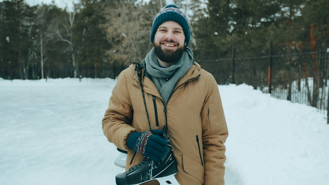 A smiling man in a warm hat and coat holding ice skates outdoors