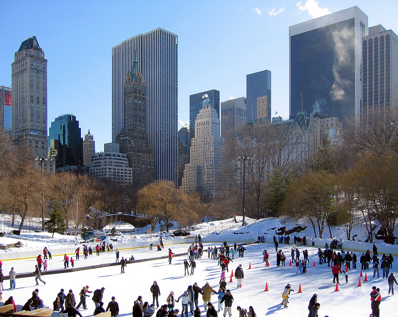 Skaters on Wollman Rink with the Manhattan skyline behind the trees